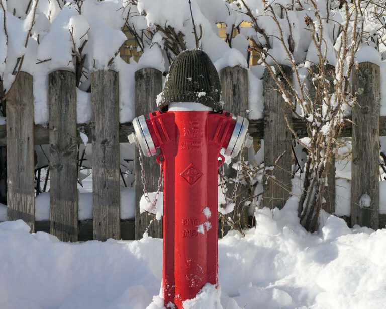 Hydrant mit Pudelmütze im zugeschneiten Clausthal-Zellerfeld vor einem hölzernen Gartenzaun.